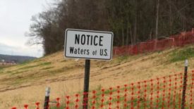 白色的长方形标志牌上写着“注意美国水域”，上面有一座棕色的小山和一排没有叶子的树