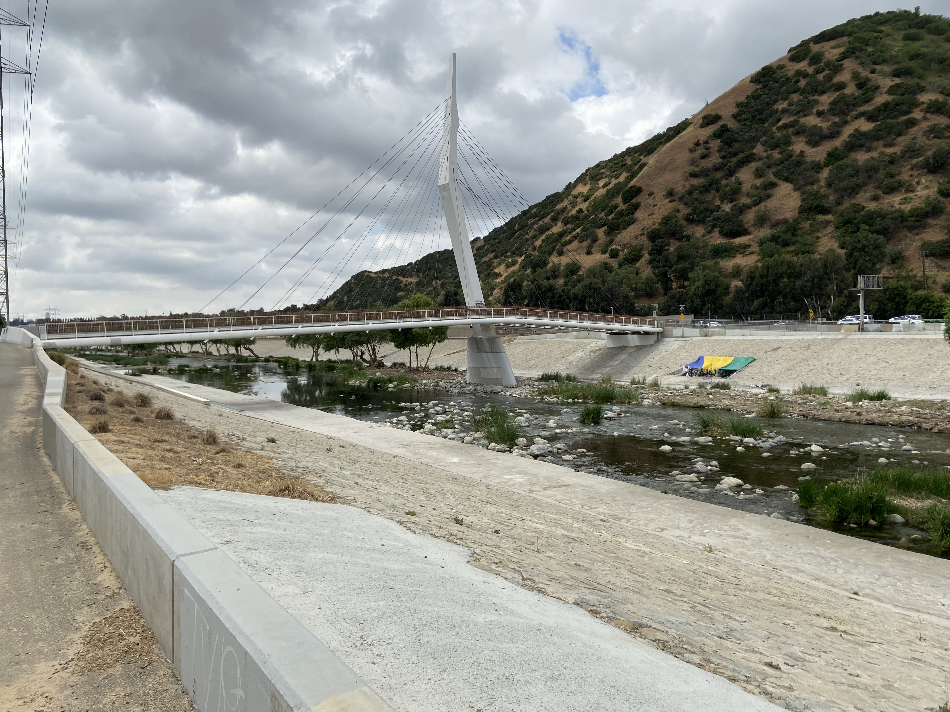A suspension bridge crosses over the LA River, encased on either side by concrete barriers. Behind the bridge is a mountain dotted with green.