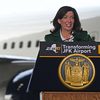 州长胡赫尔选票支持podium wearing a green suit jacket and white dress shirt. A portion of a plane at JFK airport is visible behind her.